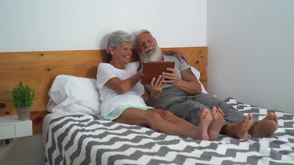 Happy senior couple using digital tablet in bed - Mature people having funny bed time together