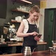 Cheerful and Content Female Barista Using Mobile Phone and Texting at Coffee Shop - VideoHive Item for Sale