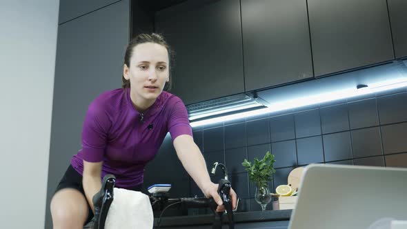 Young female drinking water from bottle while training and pedaling on stationary smart bicycle