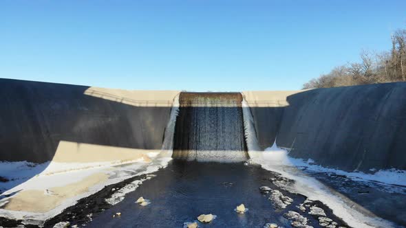 Drone flying under a bridge up and over a dam spillway out across a partially frozen lake in winter