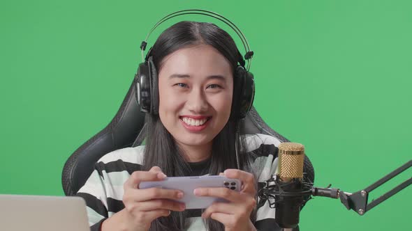 Smiling Woman With Headphone Looking At Camera While Playing Mobile Phone Game On The Green Screen