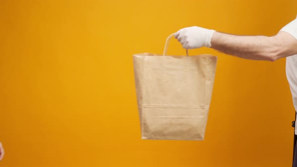 Courier in Gloves Passing Craft Shopping Bag with Delivery Against Yellow Background