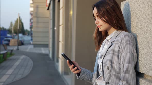 Young Woman in a Suit Looks at the Phone on the Street Near the Building