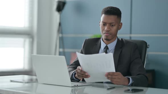 African Businessman with Laptop Reading Documents in Office