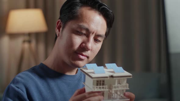 Asian Male Engineer Holding And Looking At House Model With Solar Panel While working On A Desktop