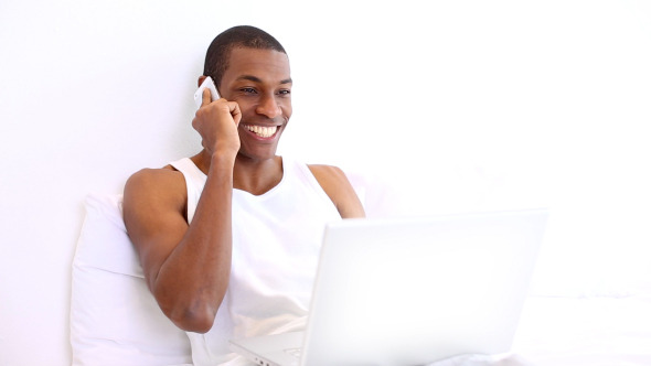 Happy Man Lying On Bed Phoning And Using Laptop