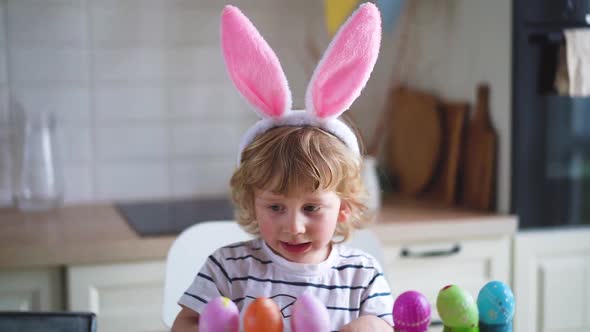 Cute two-year-old boy in bunny ears paint Easter eggs with multi-color paints sitting at the table