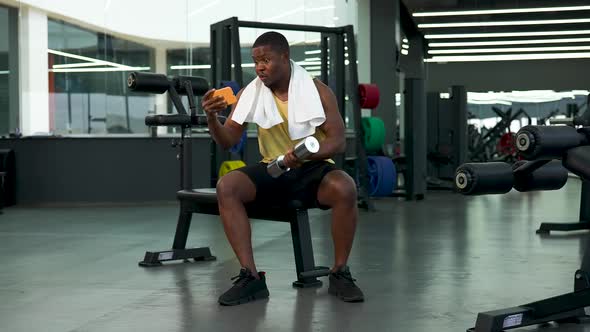 African American Man Looks at Phone in Gym While Lifting Dumbbells and Then Celebrates Victory