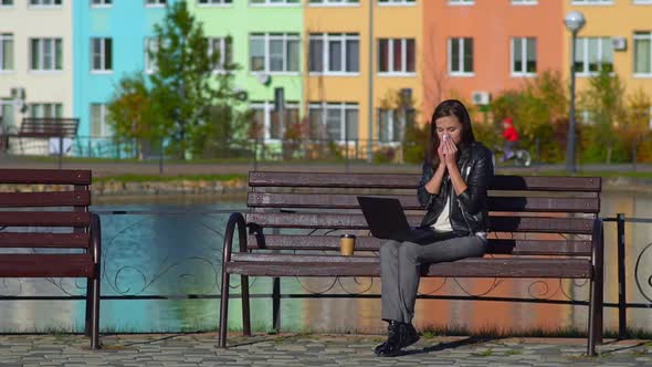 Girl in the Park on a Bench, Working in a Laptop. She Has a Cold, Wipes Her Nose with a Handkerchief