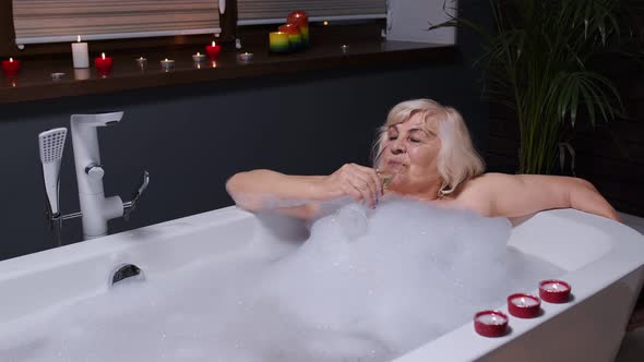 Senior Woman Grandmother Is Taking Foamy Bath, Drinking Champagne in Luxury Bathroom with Candles