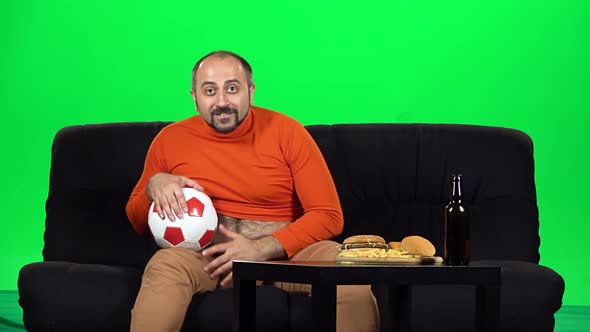 Male Football Fan Watching Soccer While Sitting on Sofa Near Table with Fast Food and Bottle of Beer