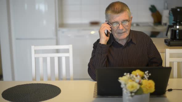 Senior Man with Eyeglasses Using Phone and Laptop in the Dining Room