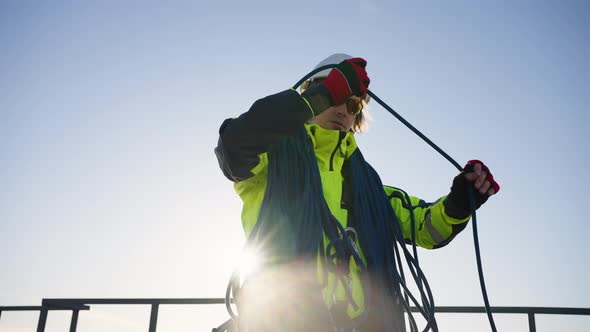 Industrial Climber in a White Helmet Coils a Rope on a Roof Against the Backdrop of the Sun
