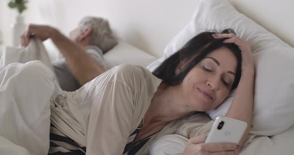 Smiling Brunette Senior Caucasian Woman Using Social Media As Mature Man Sleeping. Wife Lying in Bed