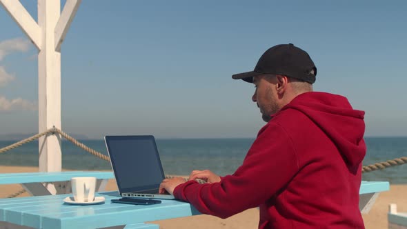 Young Man Works Remotely on a Laptop in a Cafe in a Resort