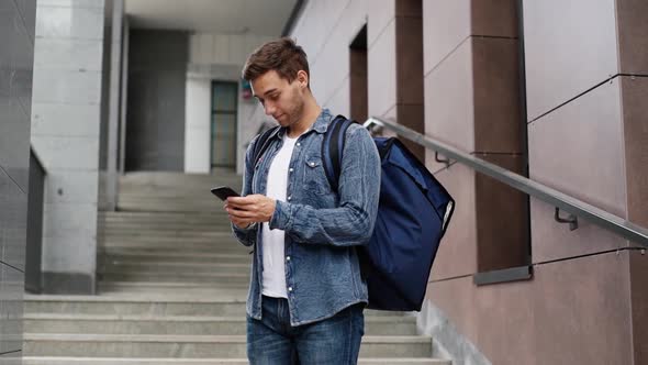 Medium Shot Portrait of Happy Delivery Man with Large Thermo Backpack Holding Phone Looking at