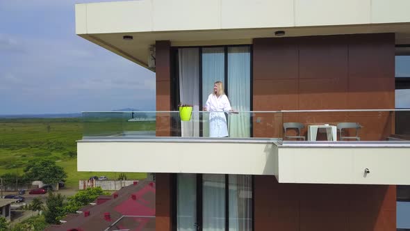 Drone view of young blonde in white coat with cup of coffee on hotel balcony