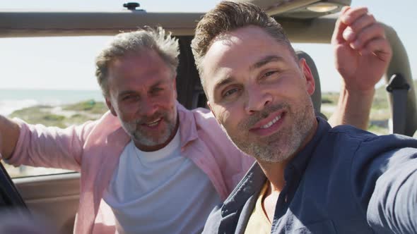 Happy caucasian gay male couple taking selfies sitting in car smiling on sunny day at the beach