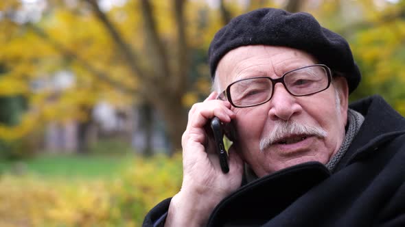 An Old Grandfather is Talking on a Smartphone He is Sitting on a Bench in the Park