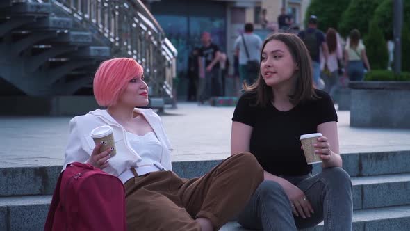 Two Young Women Talking and Laughing Sitting on the Granite Stairs and Drinking Coffee