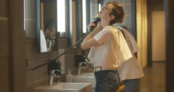 Young Woman Drying Hair with Dryer As Man Brushing Teeth at the Background. Side View of Caucasian