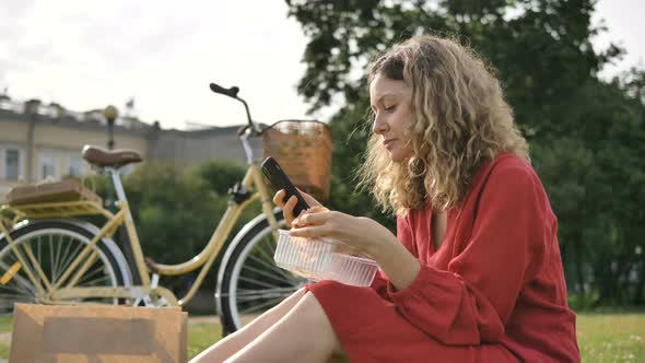 Blonde Girl in Red Dress with Phone Eats Launch in City Park