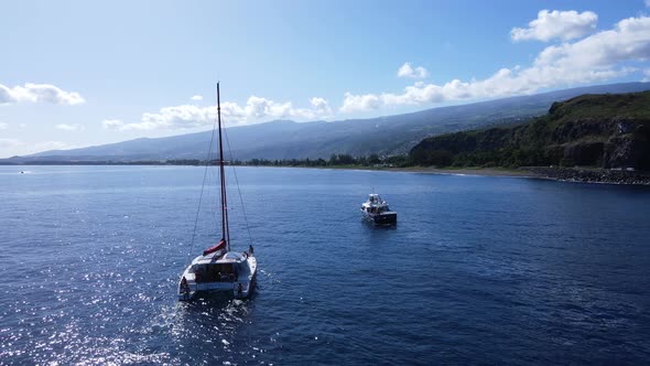 Drone footage of a catamaran with people and another boat near a cliff.