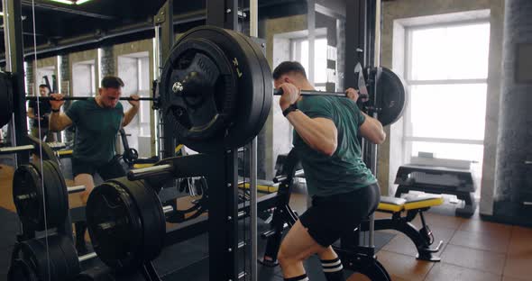 Strong Active Man In Sportswear Doing Squats Exercises With Heavy Barbell In Gym