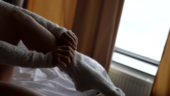 Closeup of a Woman in a Light Sweater Sitting on the Bed in the Room Pulling a White Wool Sock Over