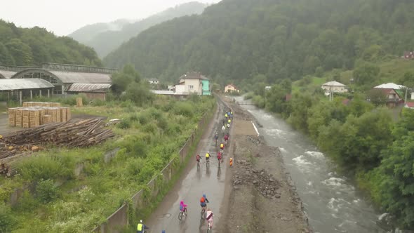 Wide aerial shot of a group of mountain bikers riding in the rain through mud alongside a river.