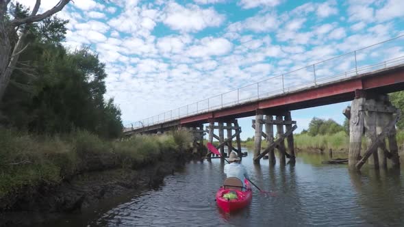 Tracking shot of a man in a red kayak paddling down a river with an old bridge extending over it.