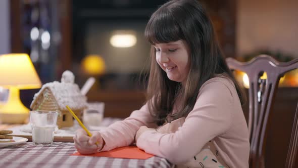 Thoughtful Pretty Caucasian Girl Writing Letter to Santa Sitting at Table at Home Indoors