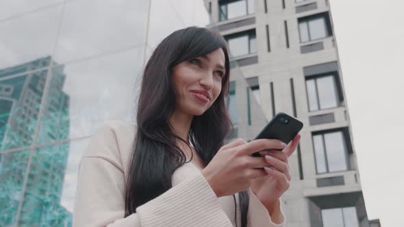 Smiling Business Woman Texting Messages on Smartphone Outdoors Near Big Modern Building. Young Woman