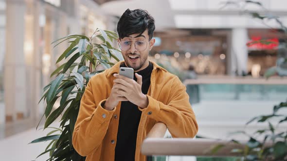 Young Hispanic Guy Stands in Mall Holding Phone Smiling Sending Email on Social Network Surfing Web