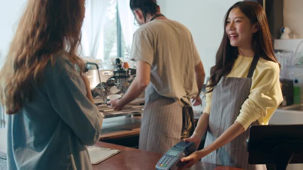 Young Asia female barista serving take away hot coffee paper cup to consumer and using credit card.