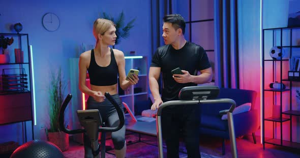 Woman and Asian Man Exercising at Home Together Using Running Track and Stationary Bike and Talking