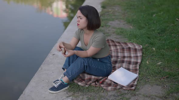 Depressed Little Woman Feeding Birds with Bread Sitting on Lake Shore in Park Thinking