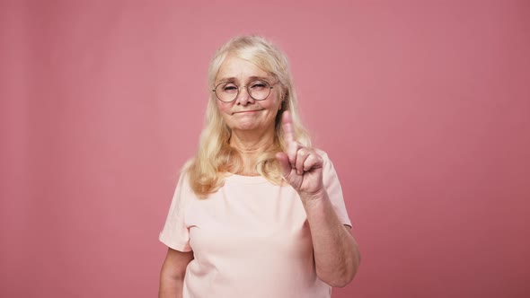 Experienced Senior Woman in Glasses Looking at Camera and Shaking Finger As Rejection Pink Studio