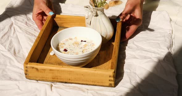 Breakfast in bed, female hands put try with bowl muesli and flower in sunlight at home