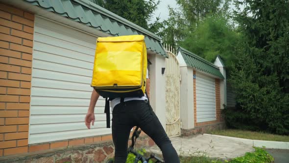 Tracking Shot of Caucasian Courier Riding Bike Walking to Fence Gate Pressing Door Bell