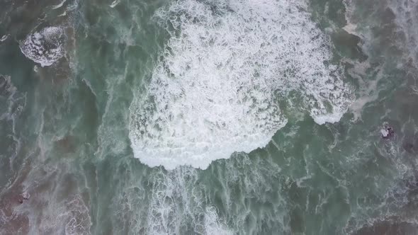 Churning white foamy waves move slowly shoreward, vertical aerial