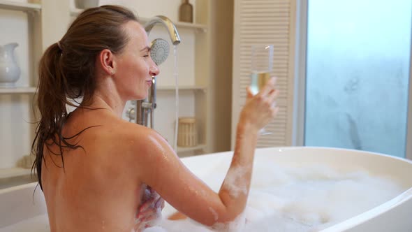 A Relaxed Woman Drinks Champagne From a Glass While Enjoying a Bubble Bath in the Bathtub