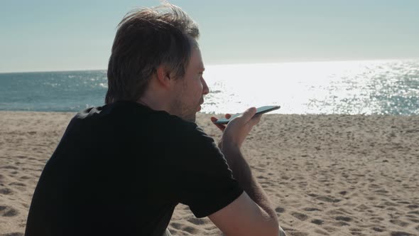 Young Man Sitting on Coastline Having Conversation with Voice Message Recorder on Smartphone