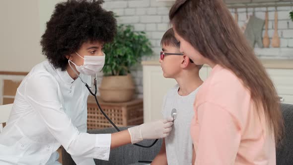 Afro American Woman Doctor in Mask Listening to Baby Breathing at Home While Sitting on Sofa