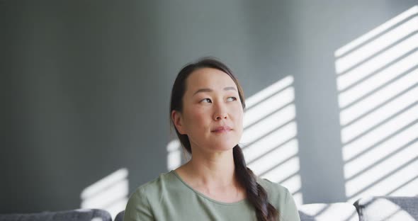 Thoughtful asian woman sitting on sofa close to window at home