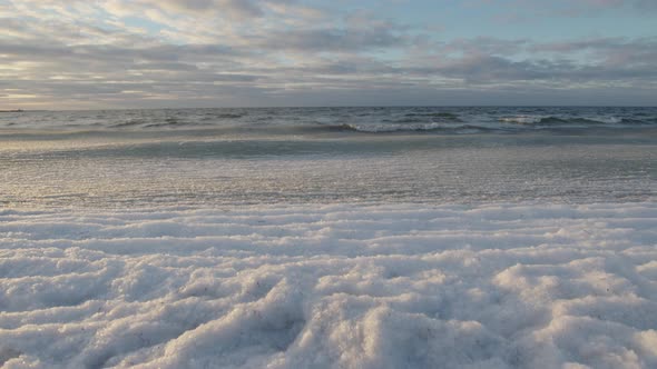 Ice covered sea waves rolling in on beach at golden hour sunset. Cloudy blue sky and white palette.