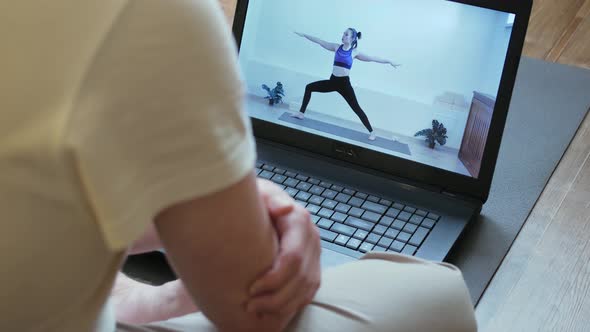 A Man Sits on a Yoga Mat at Home on Self-isolation, Watches Yoga Classes Online Using a Laptop, a