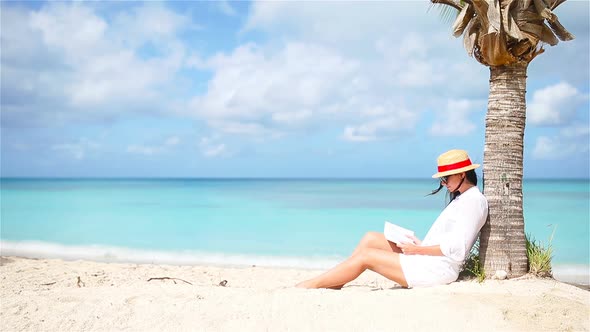Young Woman Reading Book on White Beach Sitting Under the Palmtree