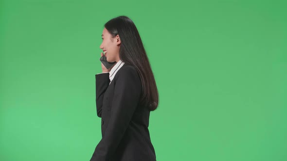 Side View Of A Smiling Asian Business Woman Talking On Mobile Phone While Walking On Green Screen