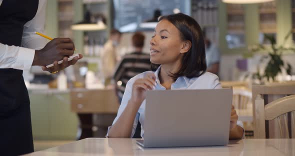 Mixedrace Businesswoman Working on Laptop and Ordering Meal in Restaurant